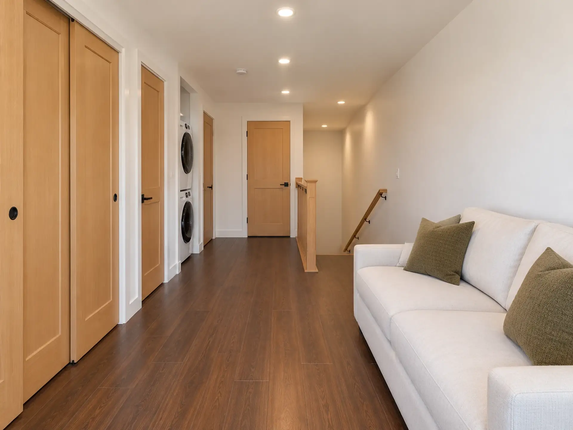 Rendering of the ADU 1200-2 loft toward the stair: on the left, a closet’s light-oak sliding doors, the door to bedroom 2, an open alcove with a stacked white washer and dryer, and the primary bedroom’s door; ahead, the linen closet’s door; on the right, the top of the stair going down along the wall beside a light-oak railing, and a white sofa with olive pillows.