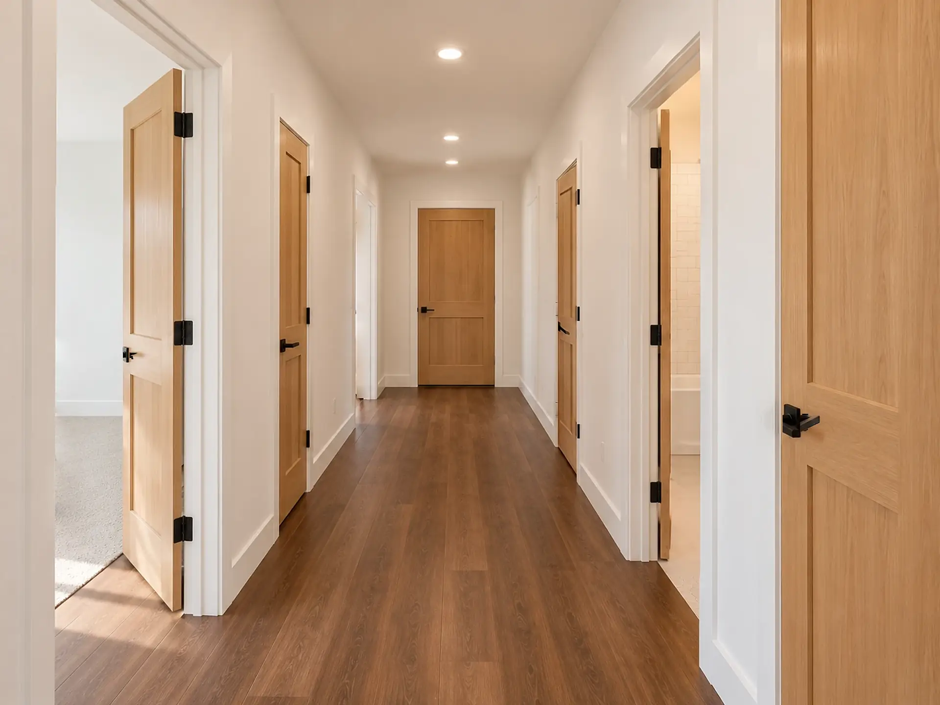 Rendering of the ADU 1200 hall toward the bedrooms: a dark plank floor between white walls, light-oak doors along both sides, an open bedroom door on the left and the hall bath open on the right, and a closed door at the far end.